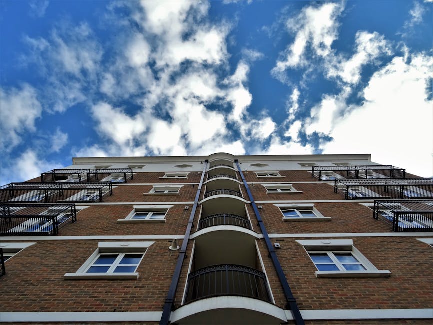 Low-angle view of a multi-storey brick residential building with black metal balcony railings and white window frames, set against a partly cloudy blue sky. The building features curved balconies on e