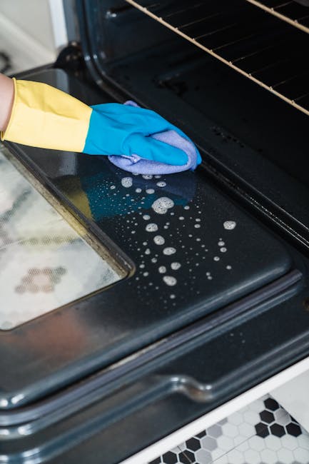 Close-up of a person wearing yellow cleaning gloves using a blue microfiber cloth to wipe the stovetop burners of a white gas range in a domestic kitchen setting. The stove surface shows signs of prev